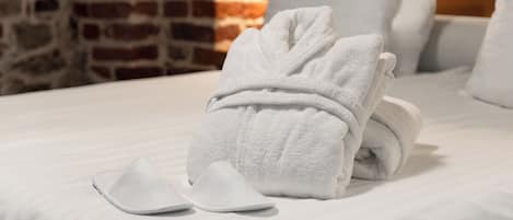 A neatly made bed with a folded towel and a pair of slippers on a white bedspread, set against a brick wall.