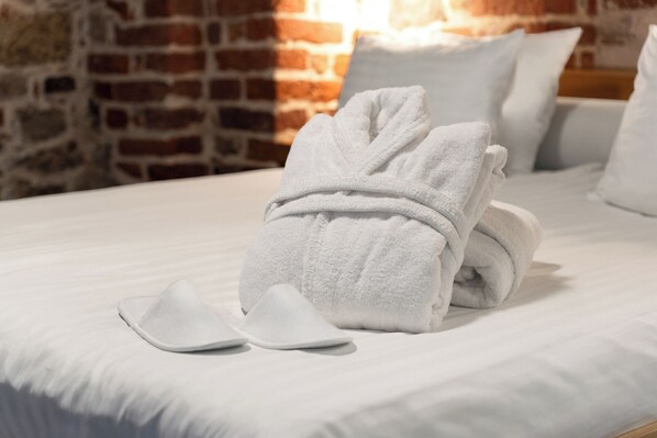 A neatly made bed with a folded towel and a pair of slippers on a white bedspread, set against a brick wall.