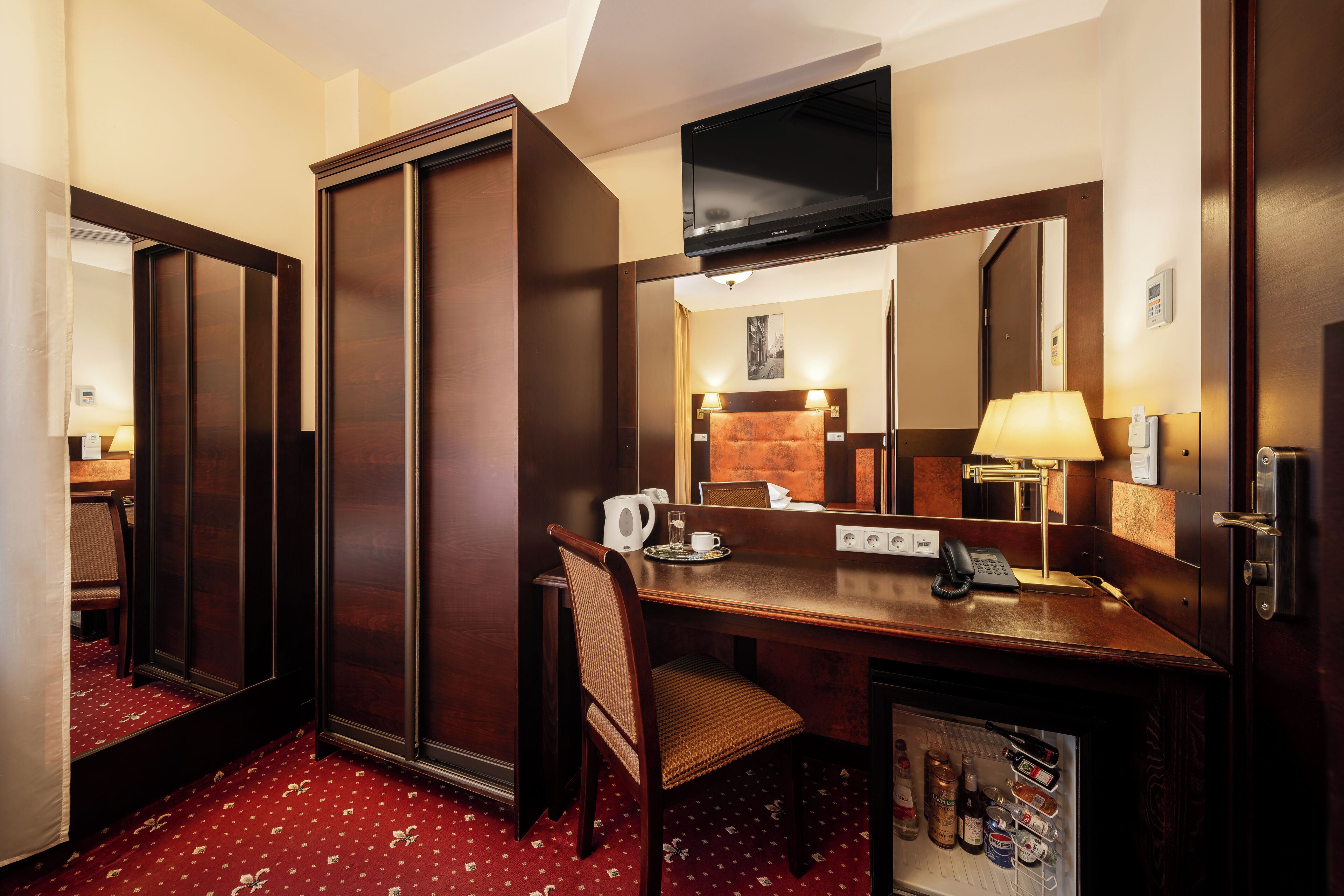 A hotel room with a wooden desk, a chair, a television mounted above the desk, a mirror, a lamp, and a minibar.