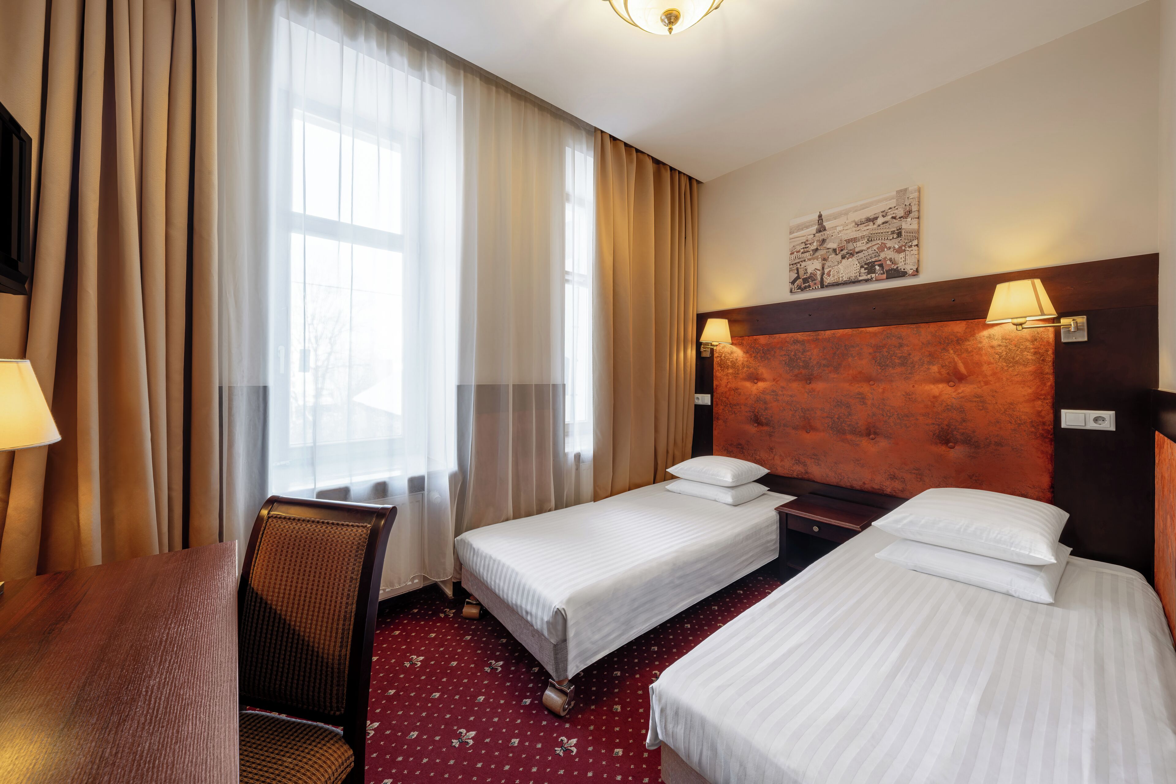 Ein Hotelzimmer mit zwei Einzelbetten, einem Holzkopfende, einem Schreibtisch, einem Stuhl und einem fenster mit Vorhängen.