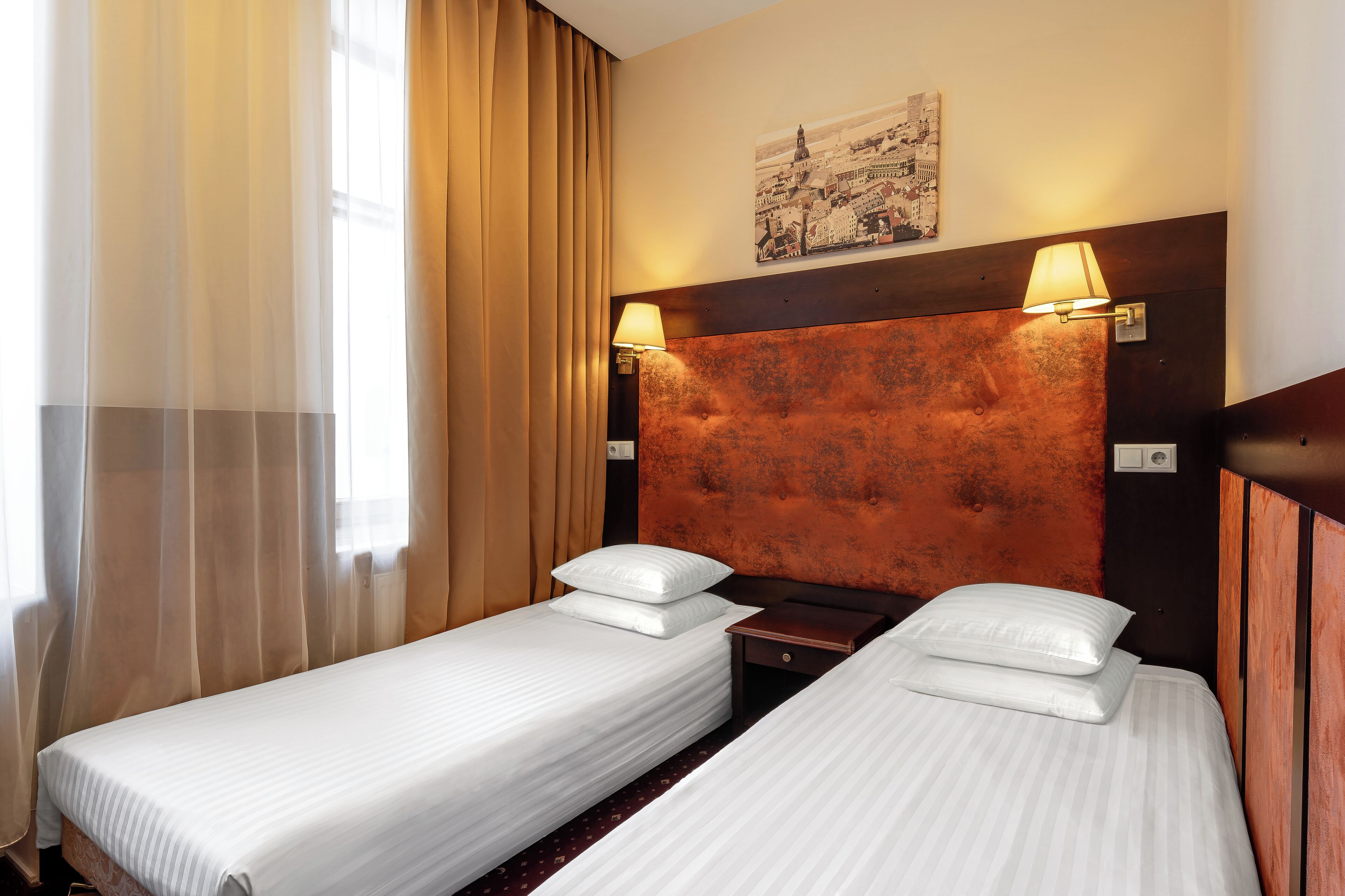 A hotel room with two single beds, a wooden headboard, a nightstand, and a wall-mounted lamp.