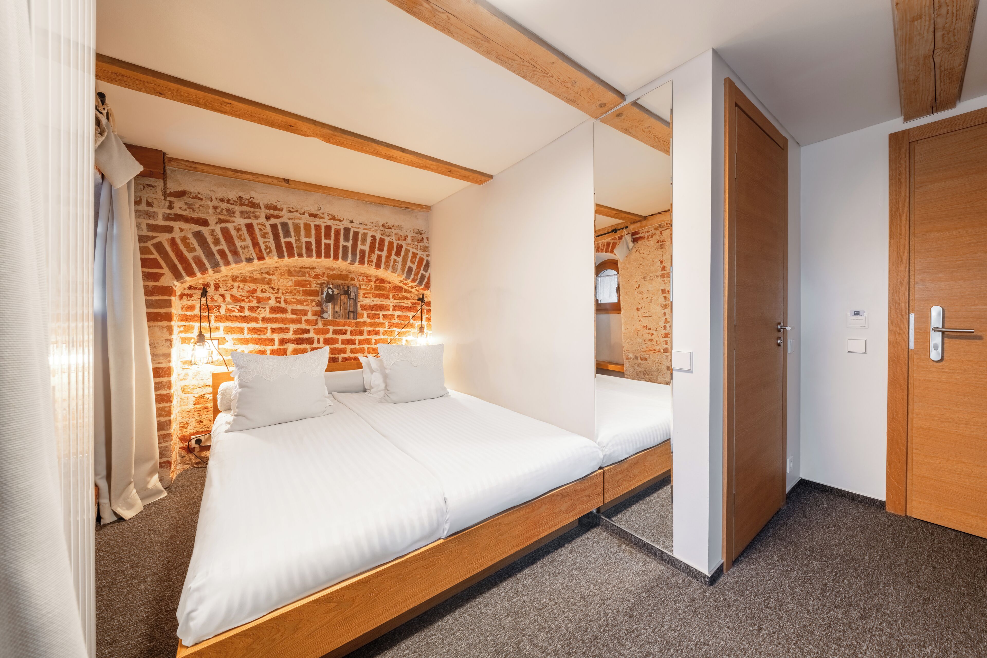 A room with a bed, a brick wall, wooden beams, and a door.