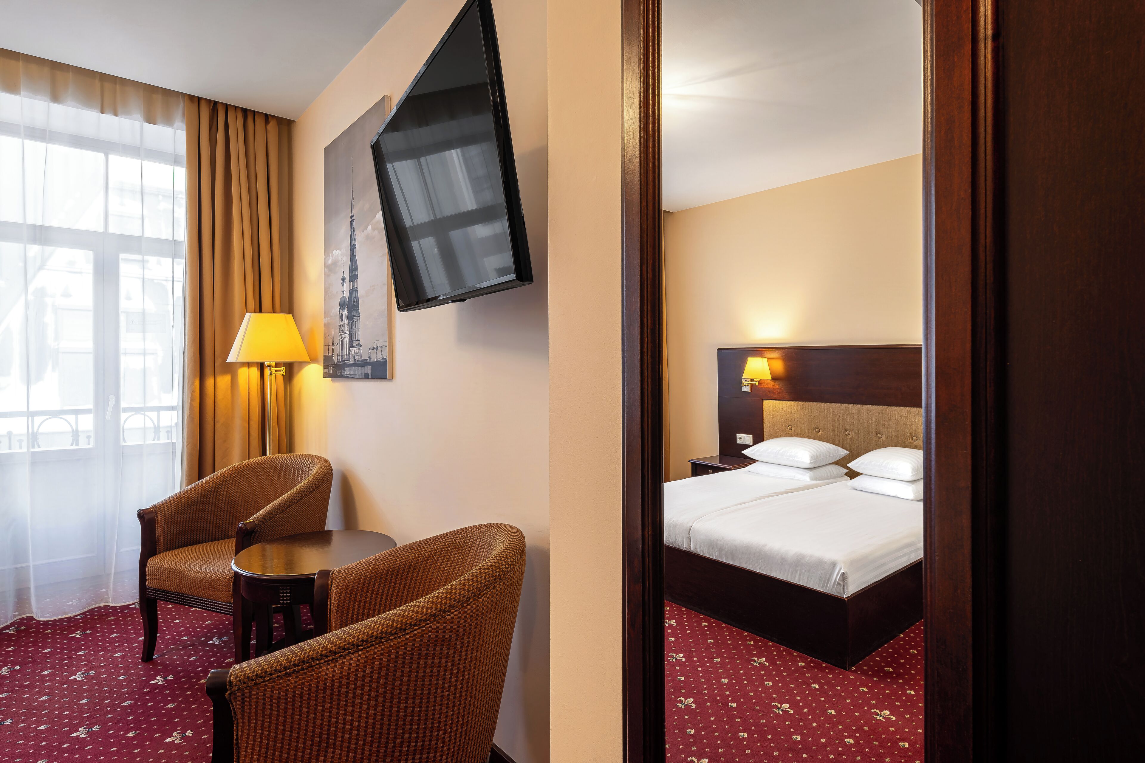 A hotel room with a bed, two chairs, a table, a TV, and a window with curtains.
