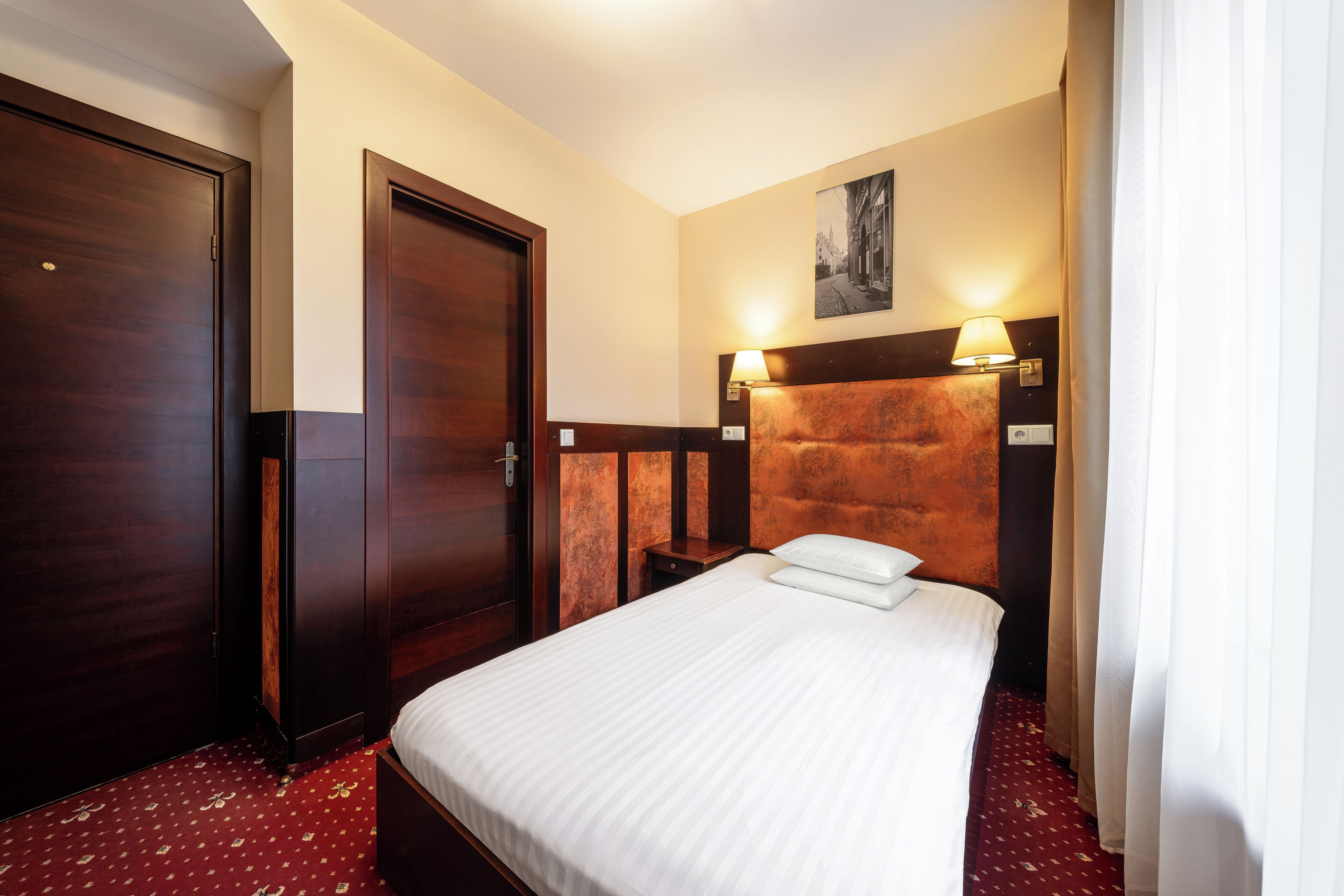 Una habitación de hotel con una cama, un armario de madera, una mesita de noche y una lámpara de pared.