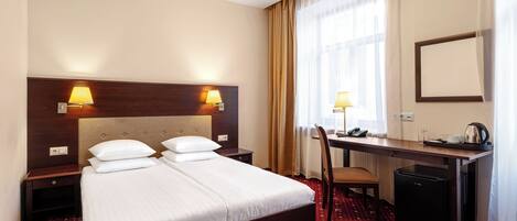 A hotel room with a double bed, bedside lamps, a desk with a chair, a refrigerator, and a large window with curtains.
