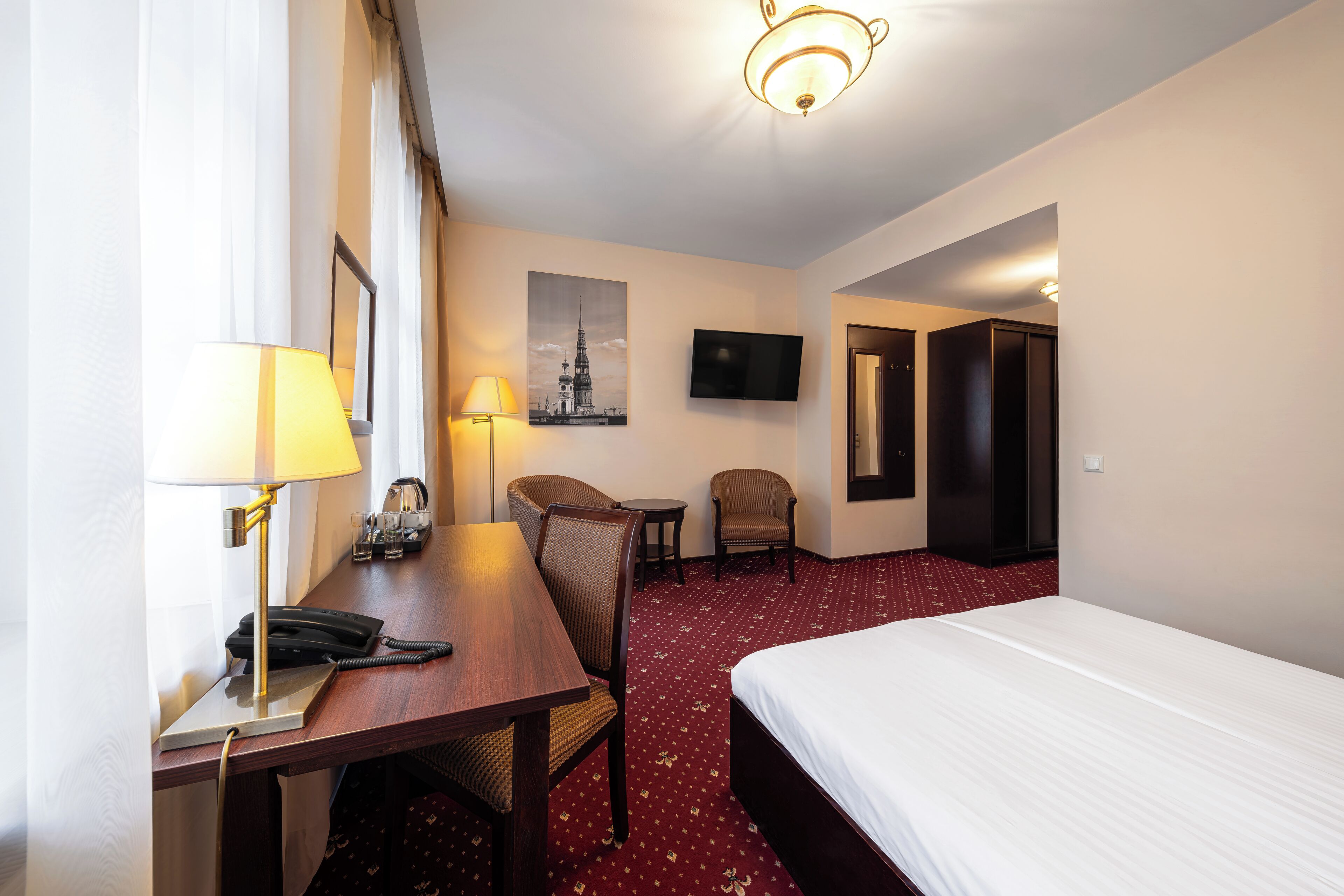 Een hotelkamer met een bed, bureau, stoelen, lamp, telefoon, televisie en een lijst met afbeelding op de muur.