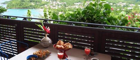 A balcony table with breakfast items, a vase of flowers, and a scenic view of a coastal town.
