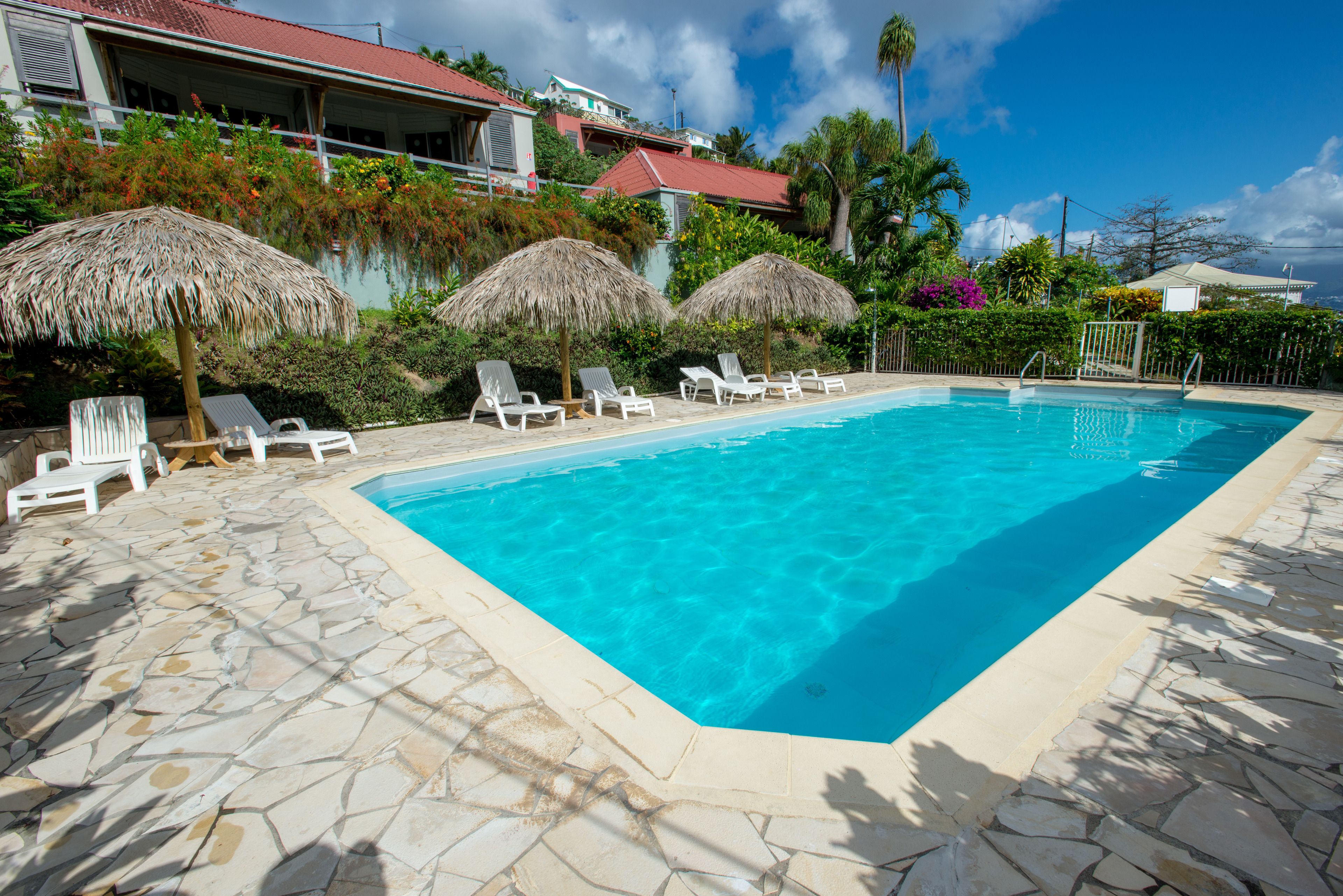 Une piscine rectangulaire à l’eau d’un bleu limpide, entourée de carreaux de pierre. Des chaises longues blanches et des parasols en chaume se trouvent le long du bord de la piscine.