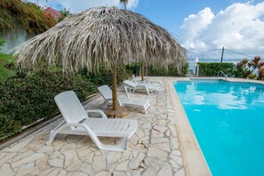 Un área junto a la piscina con sillones de descanso y una sombrilla de paja, rodeada de baldosas de piedra y follaje.