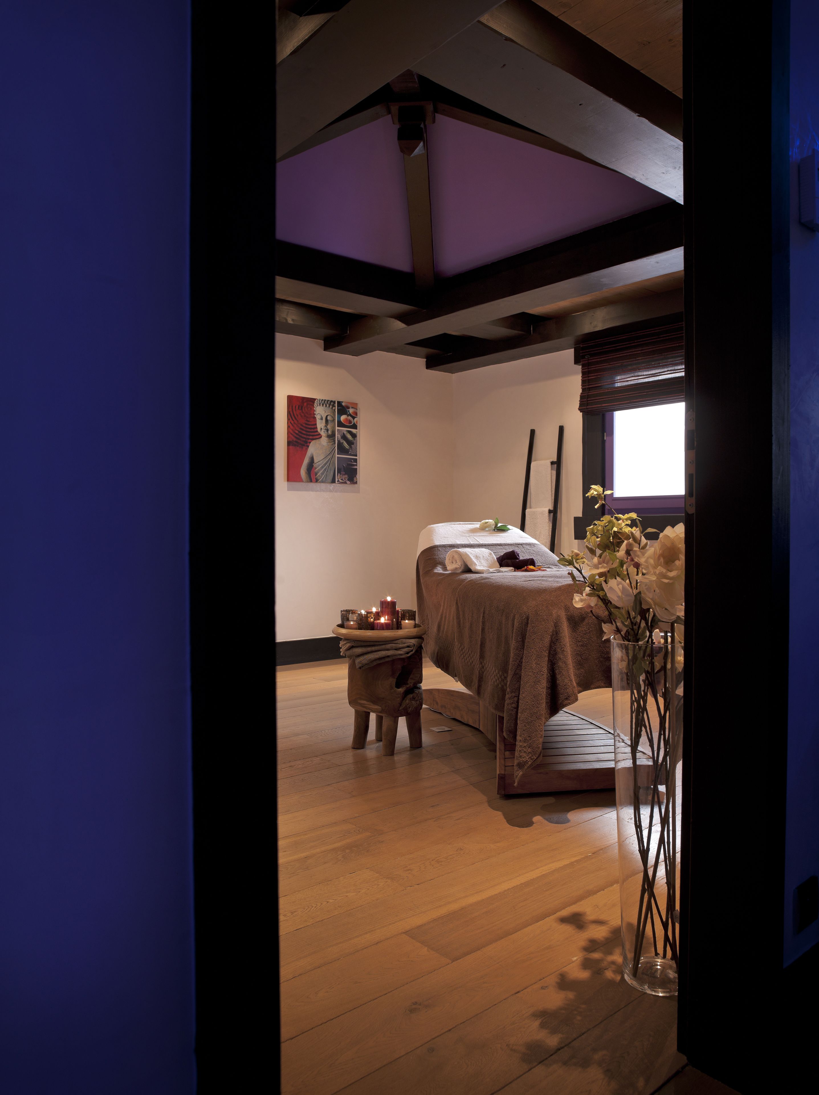 A room with a bed, a small table with candles, a vase with flowers, and a painting on the wall.
