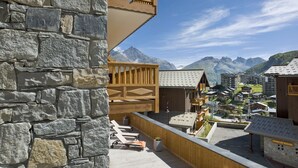 Une terrasse en bois avec des chaises longues, un mur en pierre et une vue sur les montagnes et les bâtiments.