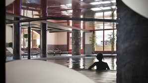 Un espace piscine intérieure où l’on voit une personne assise dans l’eau, entourée de piliers en pierre et de grandes fenêtres laissant entrer la lumière naturelle.