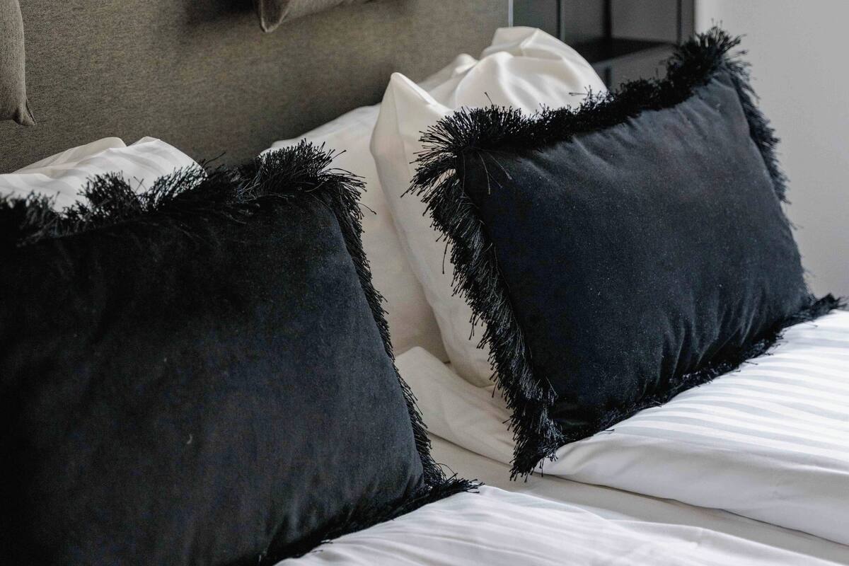 Uma cama com travesseiros brancos e escuros, uma cabeceira e uma luminária de cabeceira.