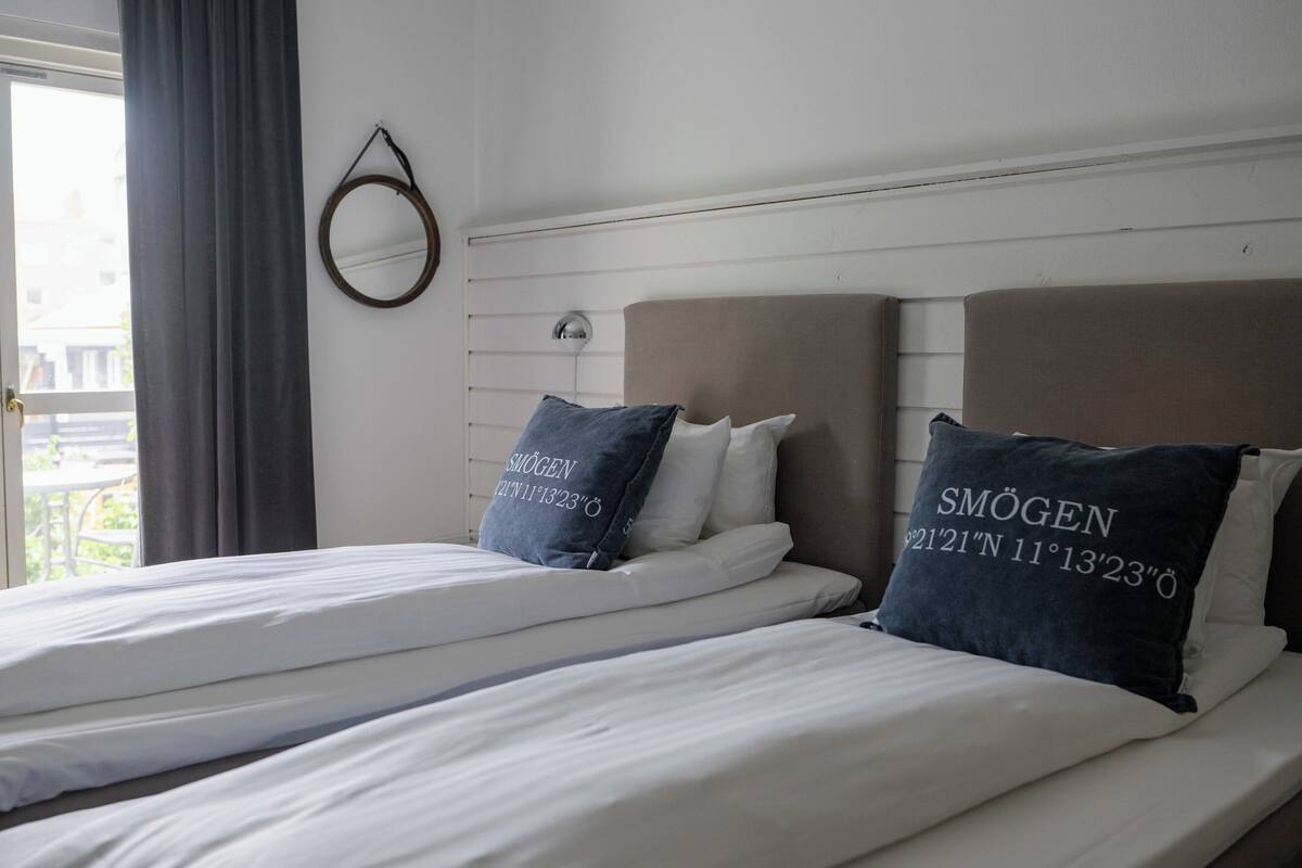 Quarto de hotel com duas camas, um espelho redondo e uma janela com cortinas.