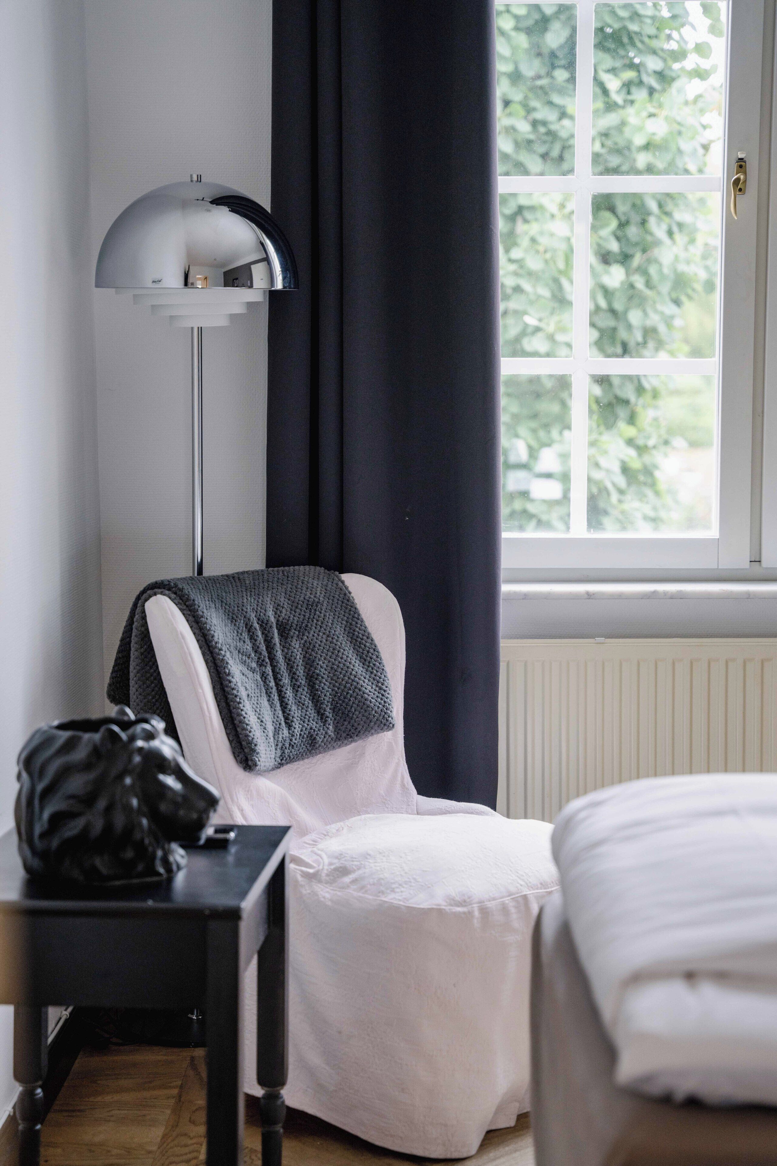 Een kamer met een bed, een stoel met plaid, een kleine tafel met tas, een vloerlamp en een raam met gordijnen.