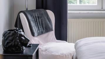 Een kamer met een bed, een stoel met plaid, een kleine tafel met tas, een vloerlamp en een raam met gordijnen.