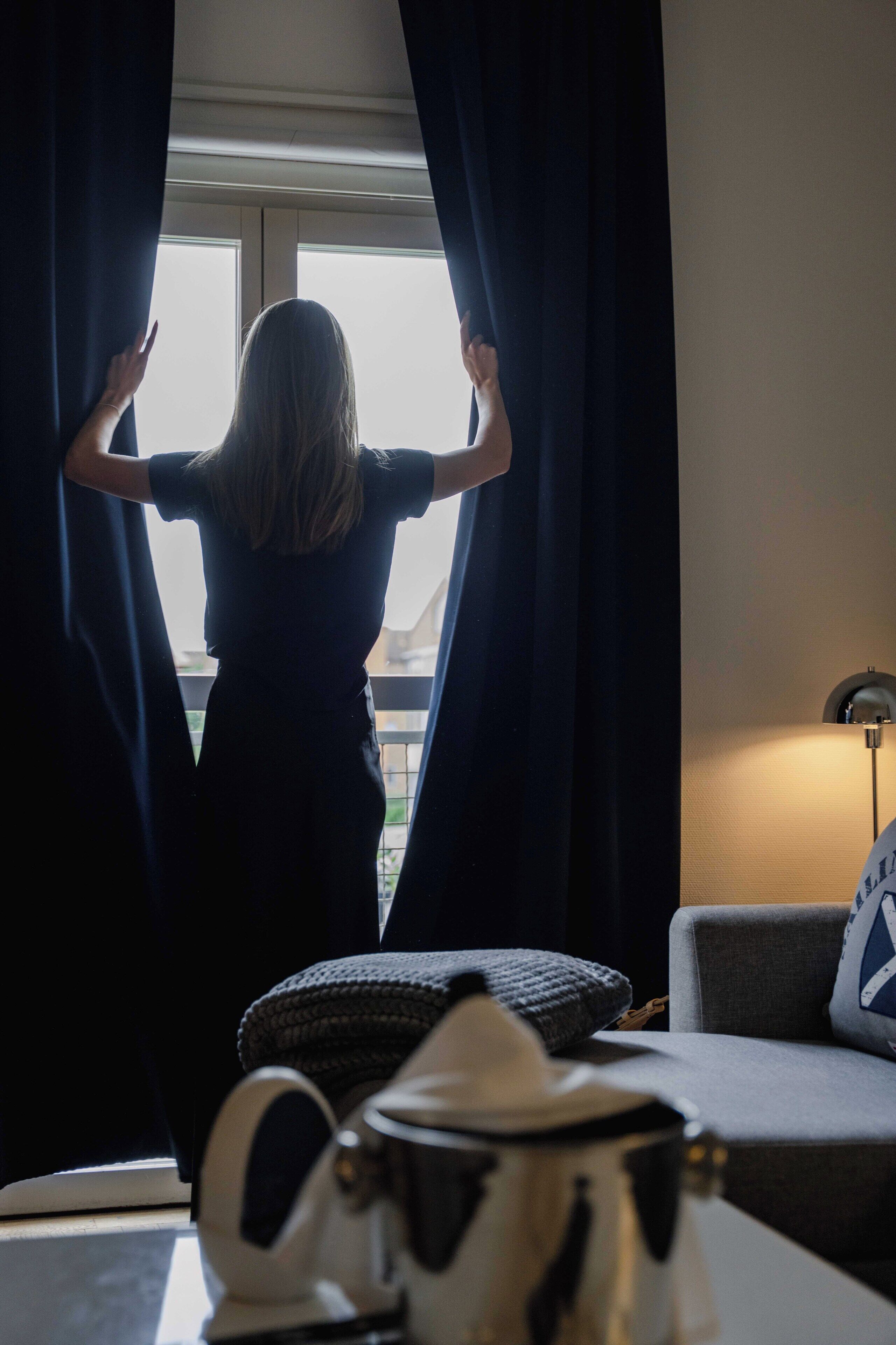 A person standing by a window with curtains, a sofa, a lamp, and a coffee pot on a table.