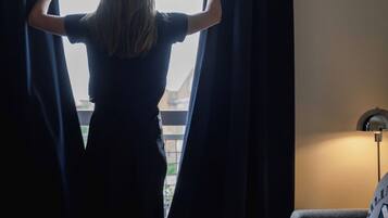 A person standing by a window with curtains, a sofa, a lamp, and a coffee pot on a table.