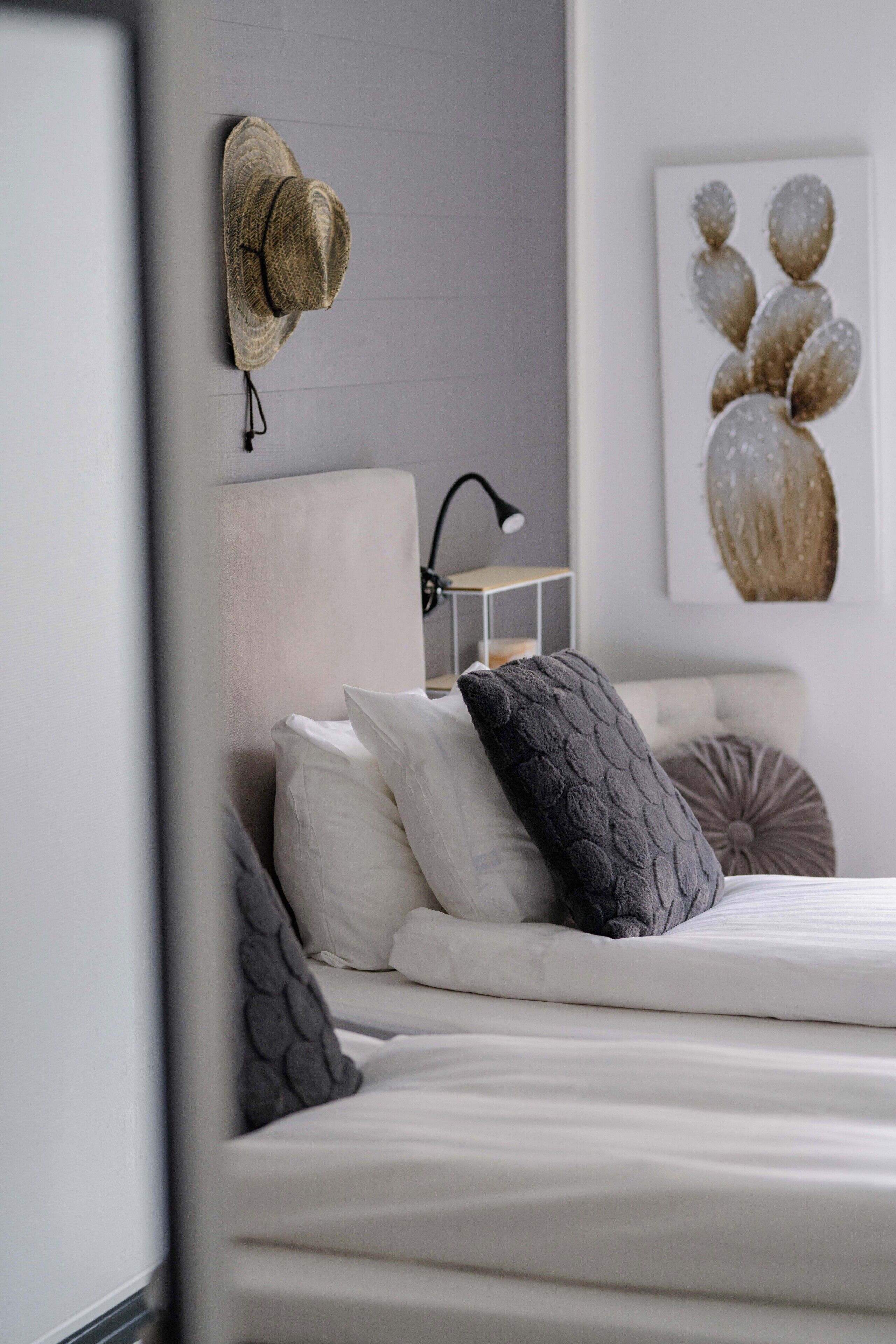 A neatly made bed with pillows, a wall-mounted hat, a lamp, and a cactus artwork.