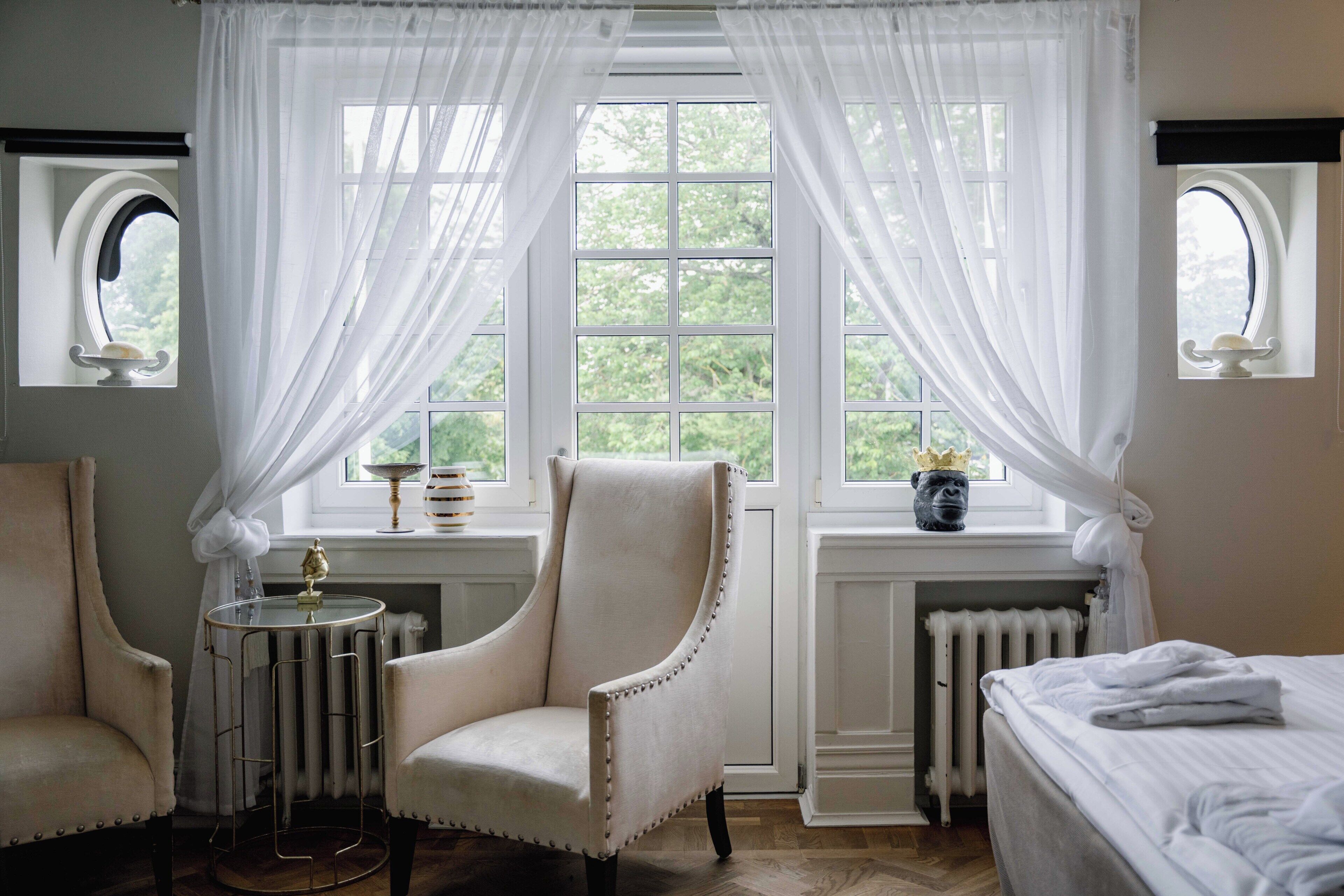A room with a large window, a radiator, two armchairs, a small table, and a bed.