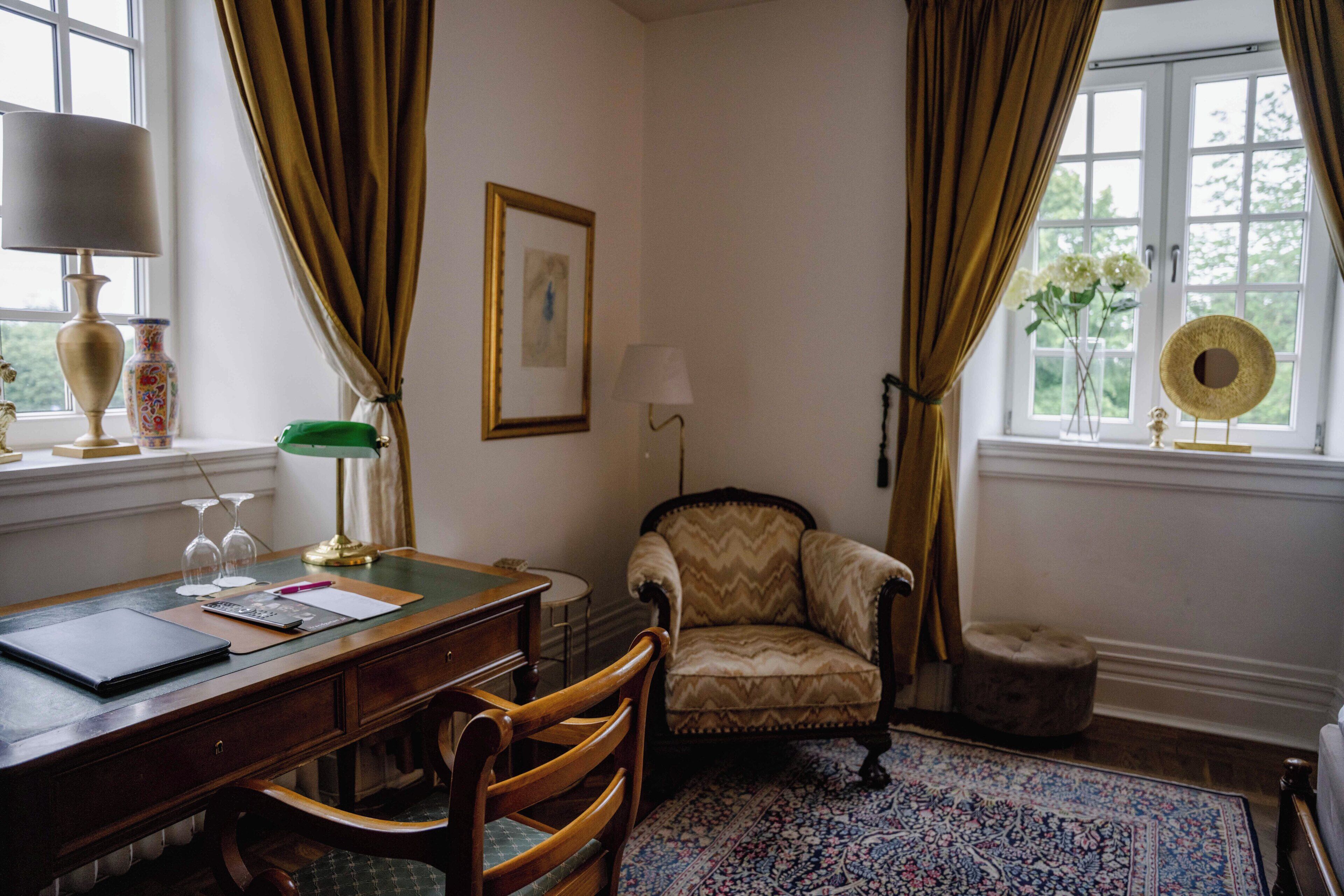 Una habitación con escritorio, silla, lámpara, cuadro enmarco y ventana con cortinas.