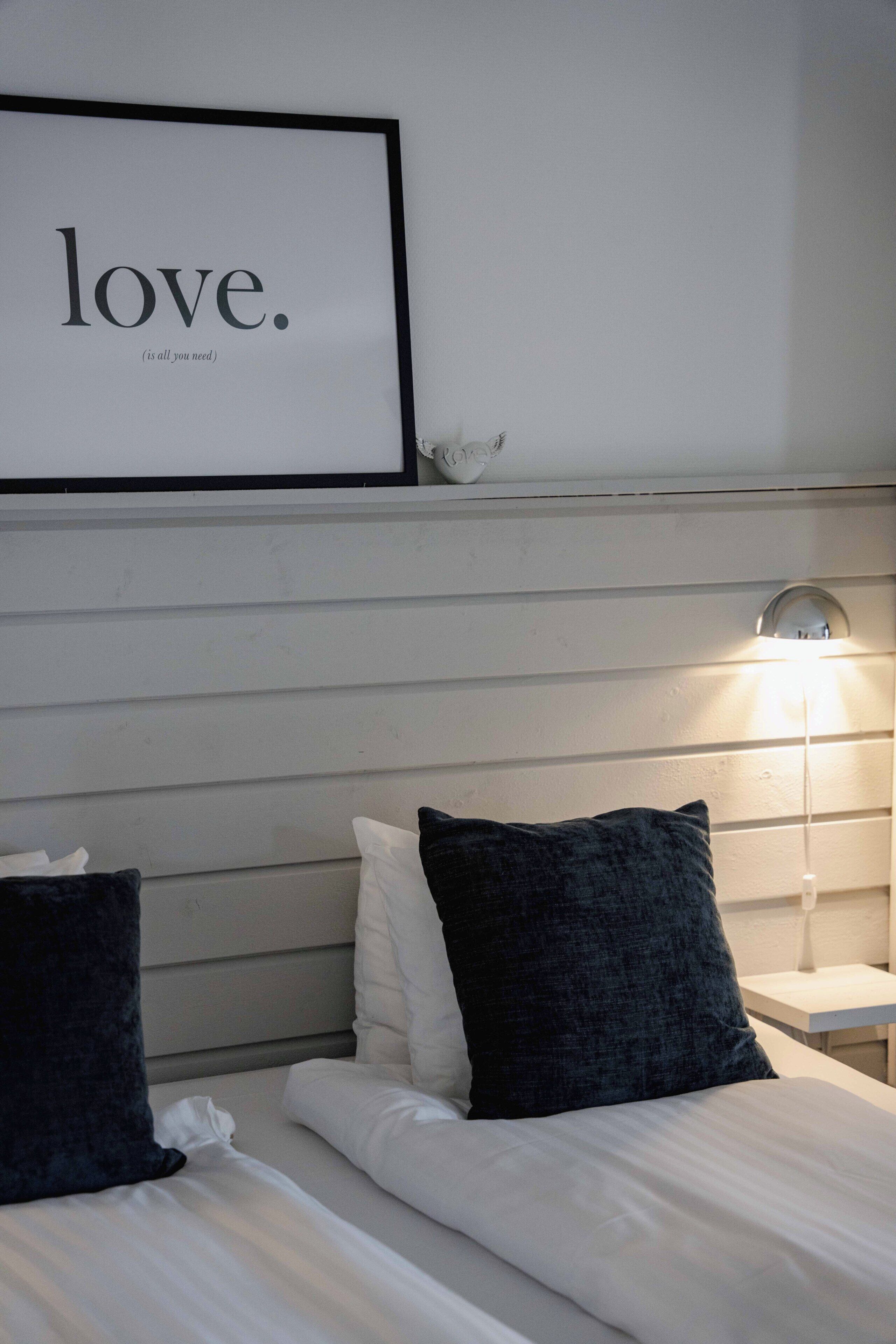 Un dormitorio con dos camas, una mesita de noche con lámpara, una cita enmarcada en la pared y un pequeño objeto decorativo en la estantería.