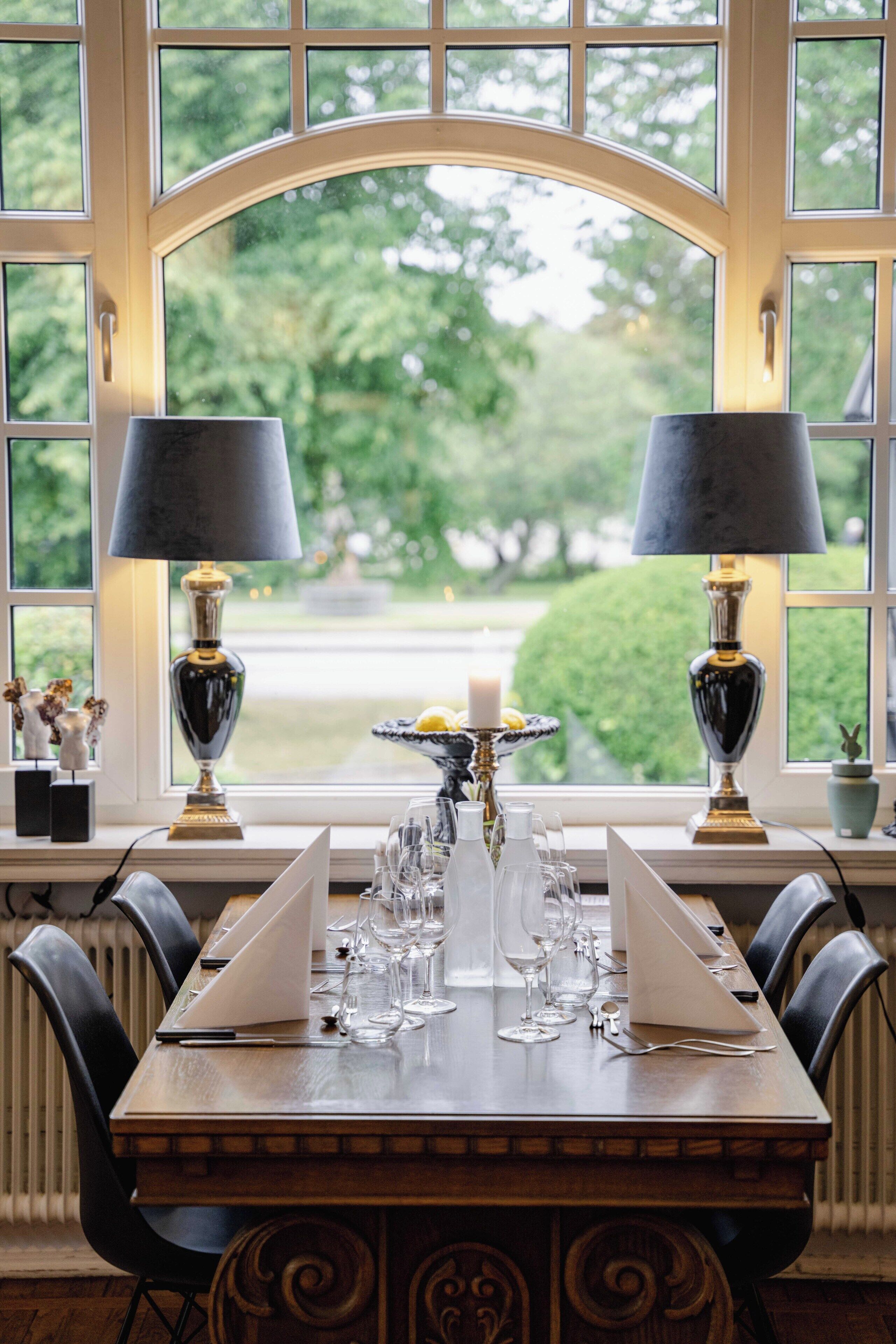 A dining table set with glasses and a candle, flanked by two lamps, in a room with a large window.