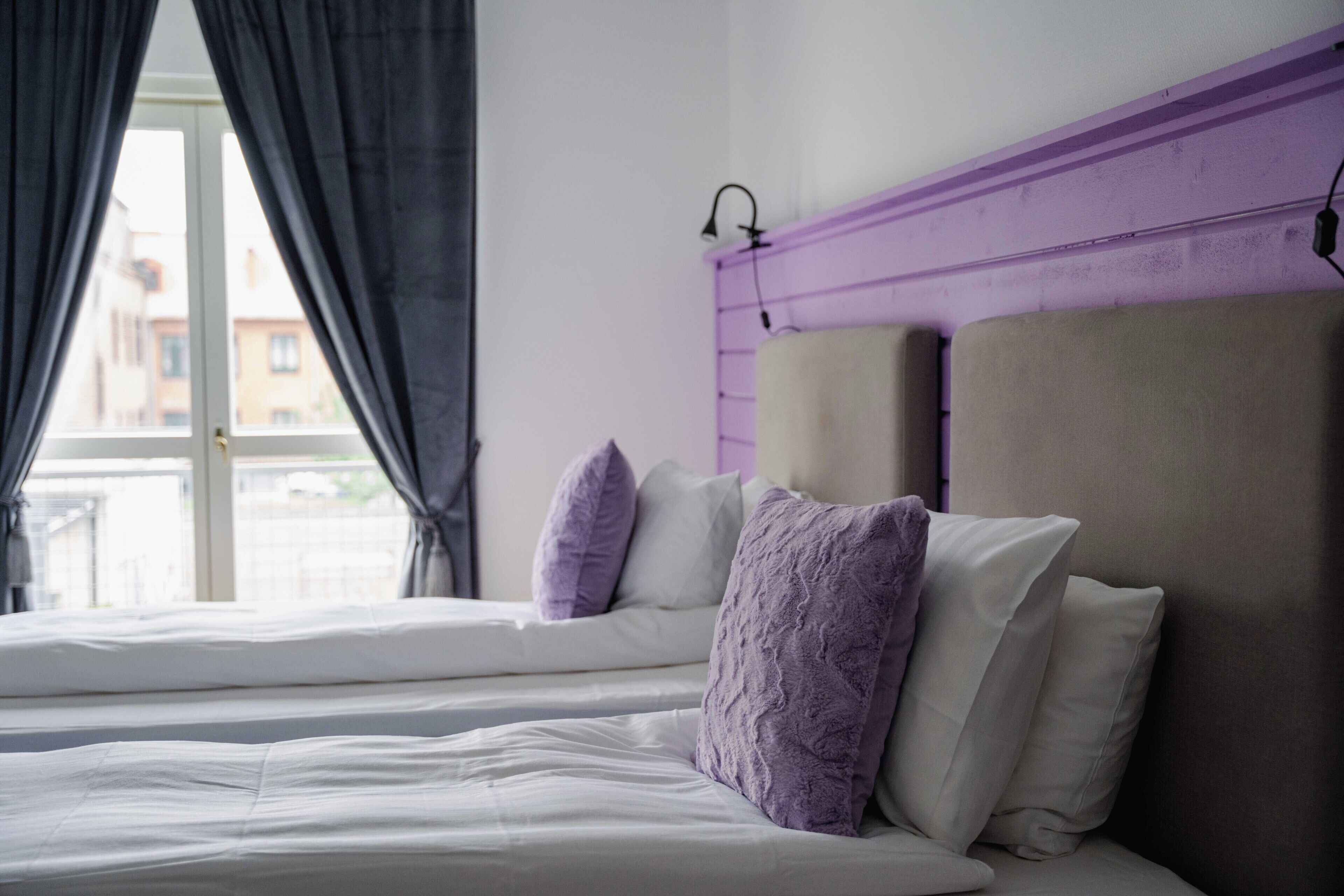 Una cama con ropa blanca y almohadas moradas, un cabecero púrpura y una ventana con cortinas.