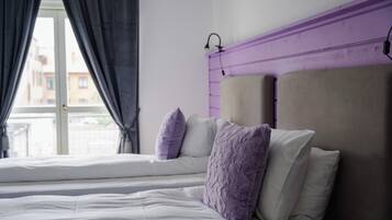 Una cama con ropa blanca y almohadas moradas, un cabecero púrpura y una ventana con cortinas.