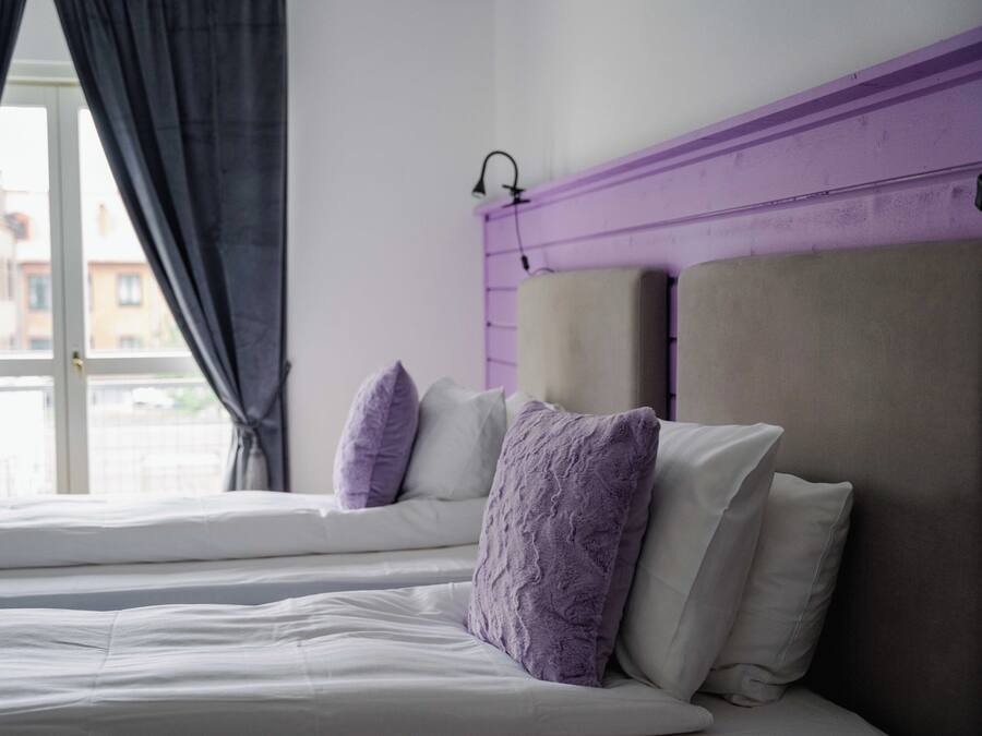Una cama con ropa blanca y almohadas moradas, un cabecero púrpura y una ventana con cortinas.
