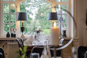 Mesa de comedor para dos personas, con dos lámparas, velas y vistas a follaje a través de ventanas amplias.