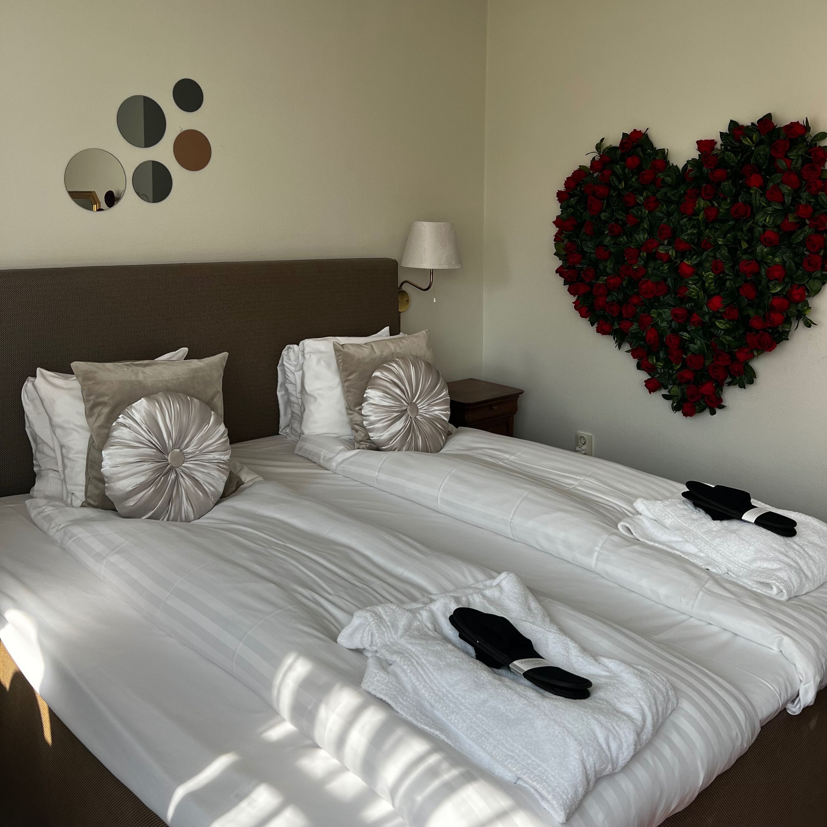 Una cama bien hecha con sábanas blancas, un arreglo floral con forma de corazón en la pared y una mesita de noche con lámpara.