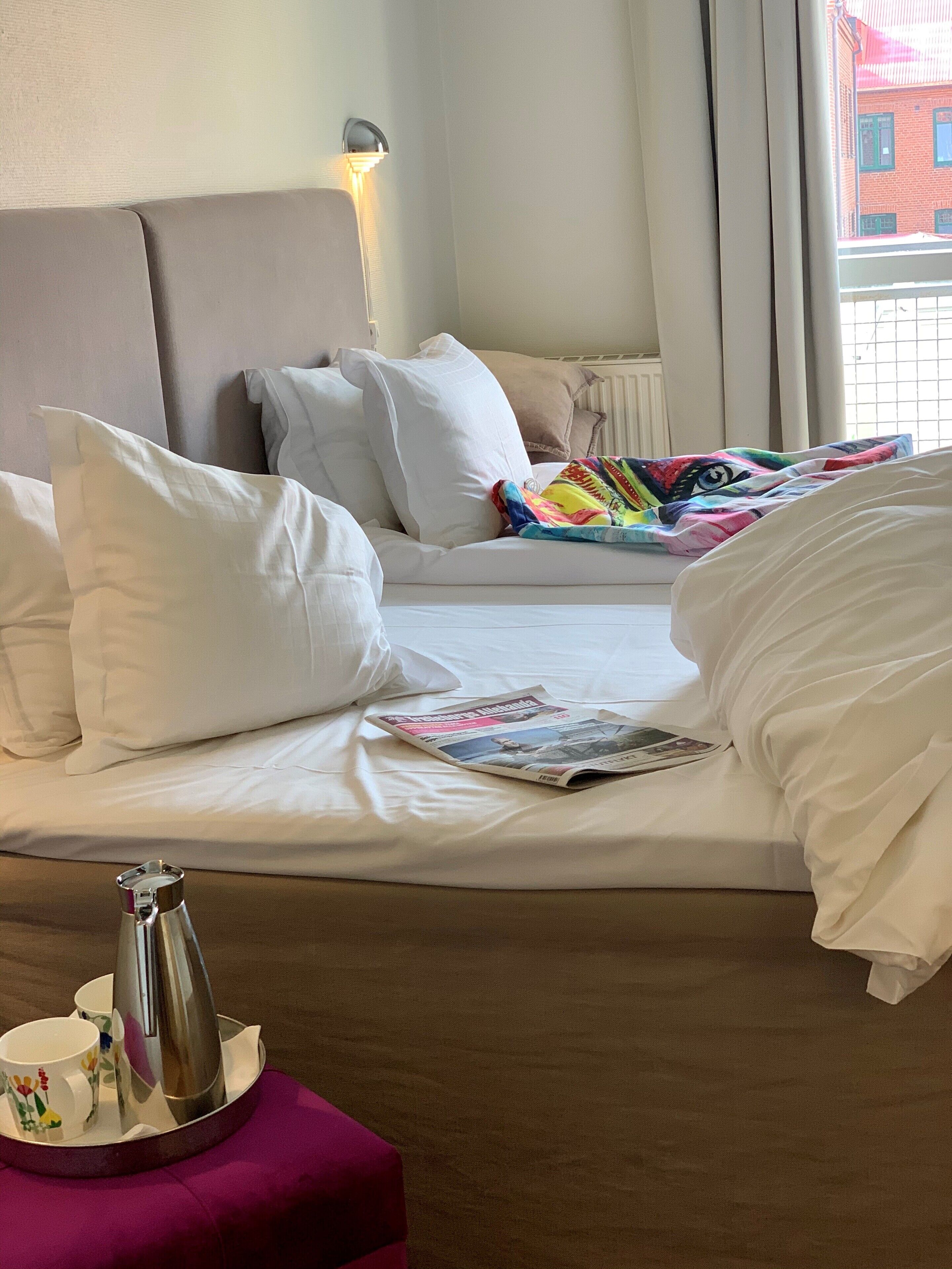 A hotel room with a bed, pillows, a magazine, a tray with a teapot and cups, and a window with a view of buildings.