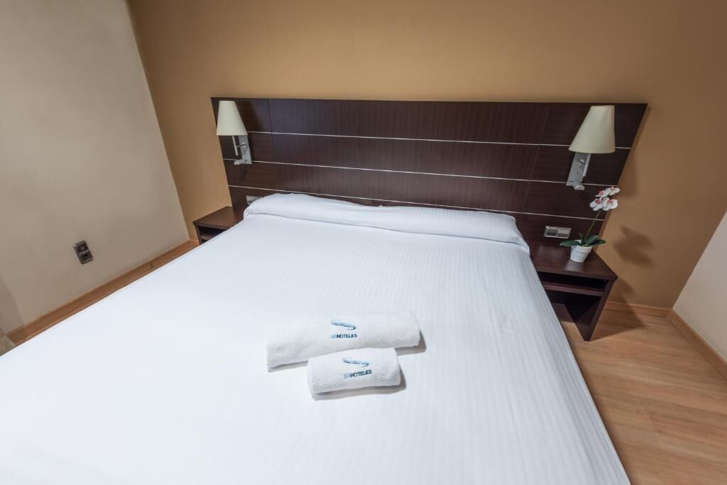 A hotel room with a large bed, two pillows, two white towels, a nightstand with a lamp, and a small table with a potted plant.