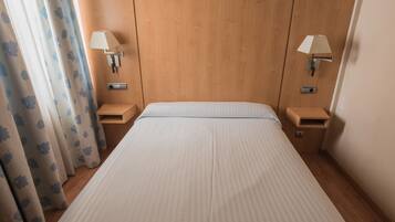 Una cama bien tendida con cabecera de madera, dos mesitas de noche con lámparas y una cortina con estampado floral.
