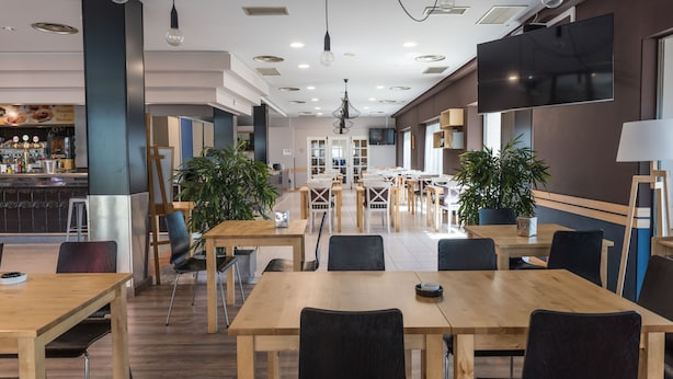 A modern dining area with wooden tables and chairs, potted plants, and a bar in the background. - Hotel Las Ventas (Utebo)