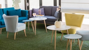 A modern lounge area with a blue sofa, a grey armchair, and a yellow armchair. There are two round tables with drinks and a small plant. - Hotel Las Ventas (Utebo)