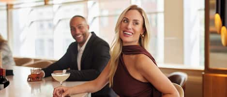 A person in a dark dress seated at a bar with a martini glass, another person in a suit in the background, and a modern interior with a chandelier.