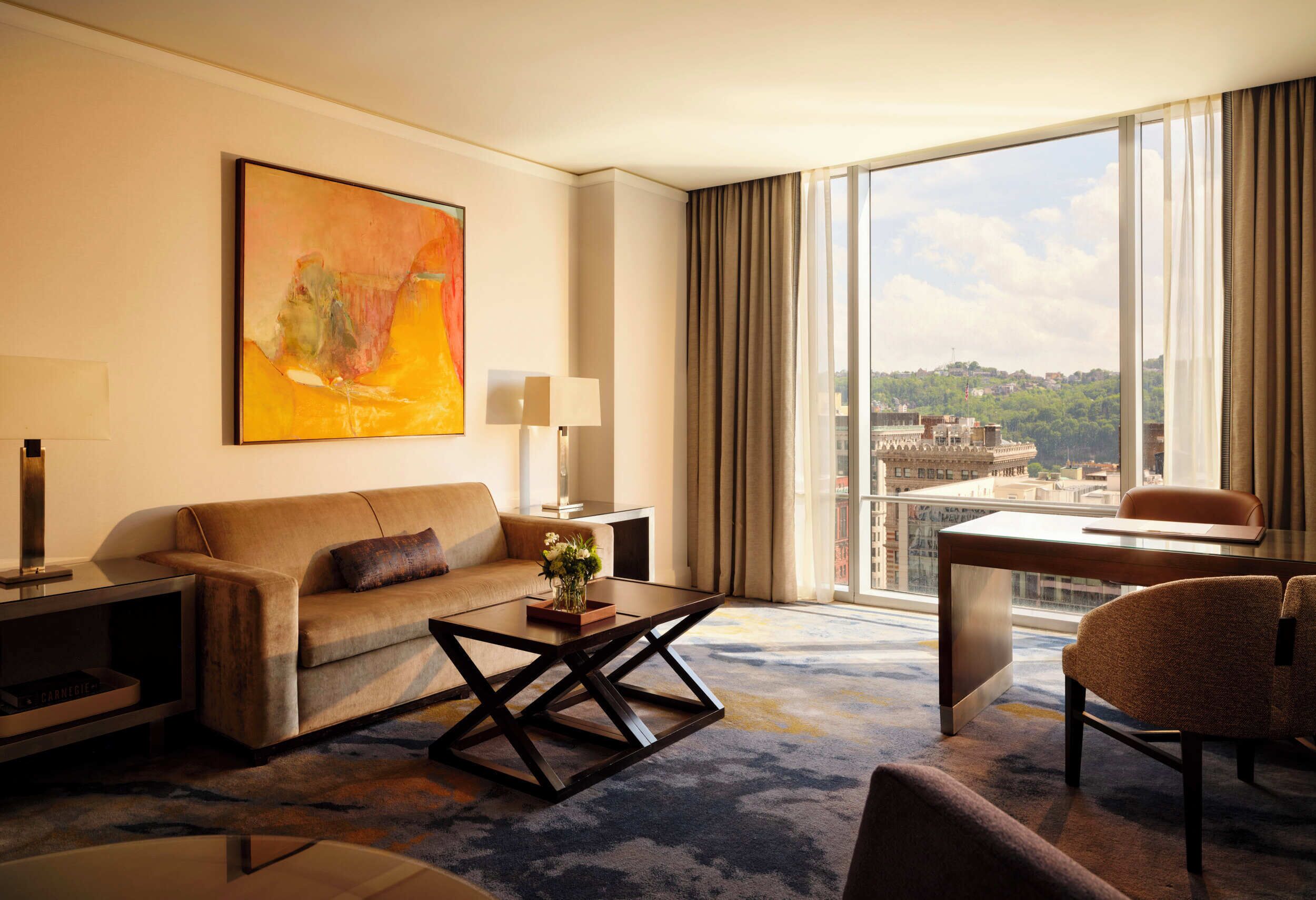 A hotel room with a sofa, coffee table, and a desk with a chair. There is a large window with a view of buildings and a painting on the wall.