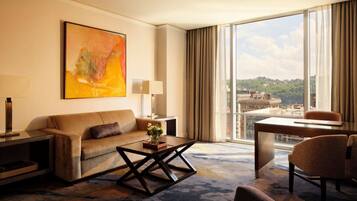 A hotel room with a sofa, coffee table, and a desk with a chair. There is a large window with a view of buildings and a painting on the wall.