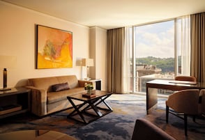 Habitación de hotel con sofá, mesa de centro y escritorio con silla. Amplia ventana con vistas a edificios y un cuadro en la pared.