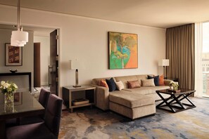 A modern hotel room with a sofa, a coffee table, a dining table with chairs, a painting on the wall, and a view of the city through the window.
