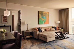 Una habitación de hotel moderna con sofá, una mesa de centro, una mesa de comedor con sillas, un cuadro en la pared y vistas a la ciudad a través del ventanal.