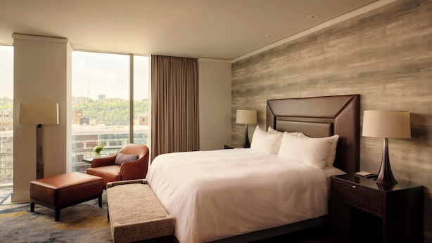 A hotel room with a large bed, a chair, a bench, a nightstand, and a view of the city. - Fairmont Pittsburgh (Pittsburgh)