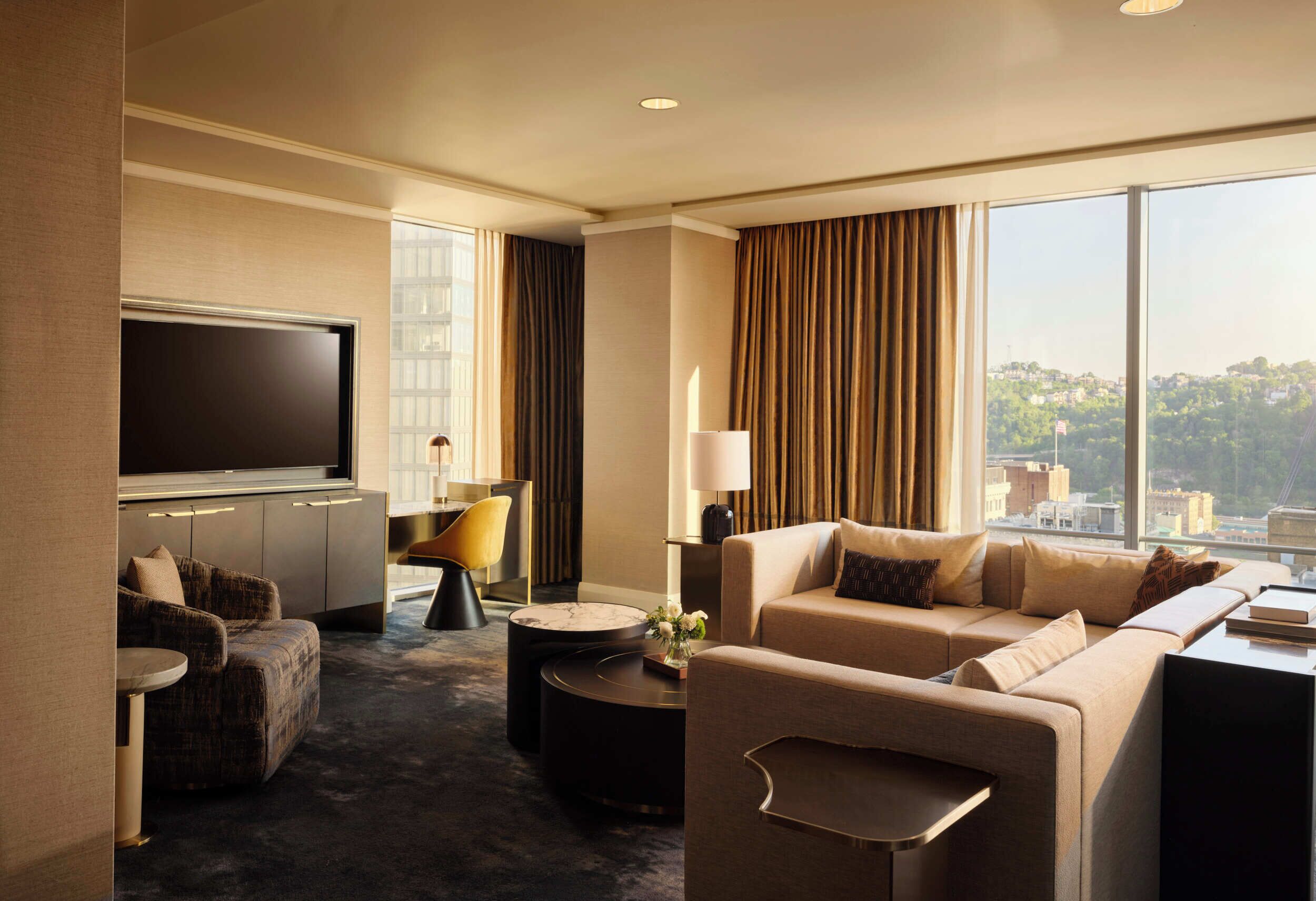 Habitación de hotel con televisor de pantalla plana grande, un sofá, una silla, una mesita y vista a la ciudad.
