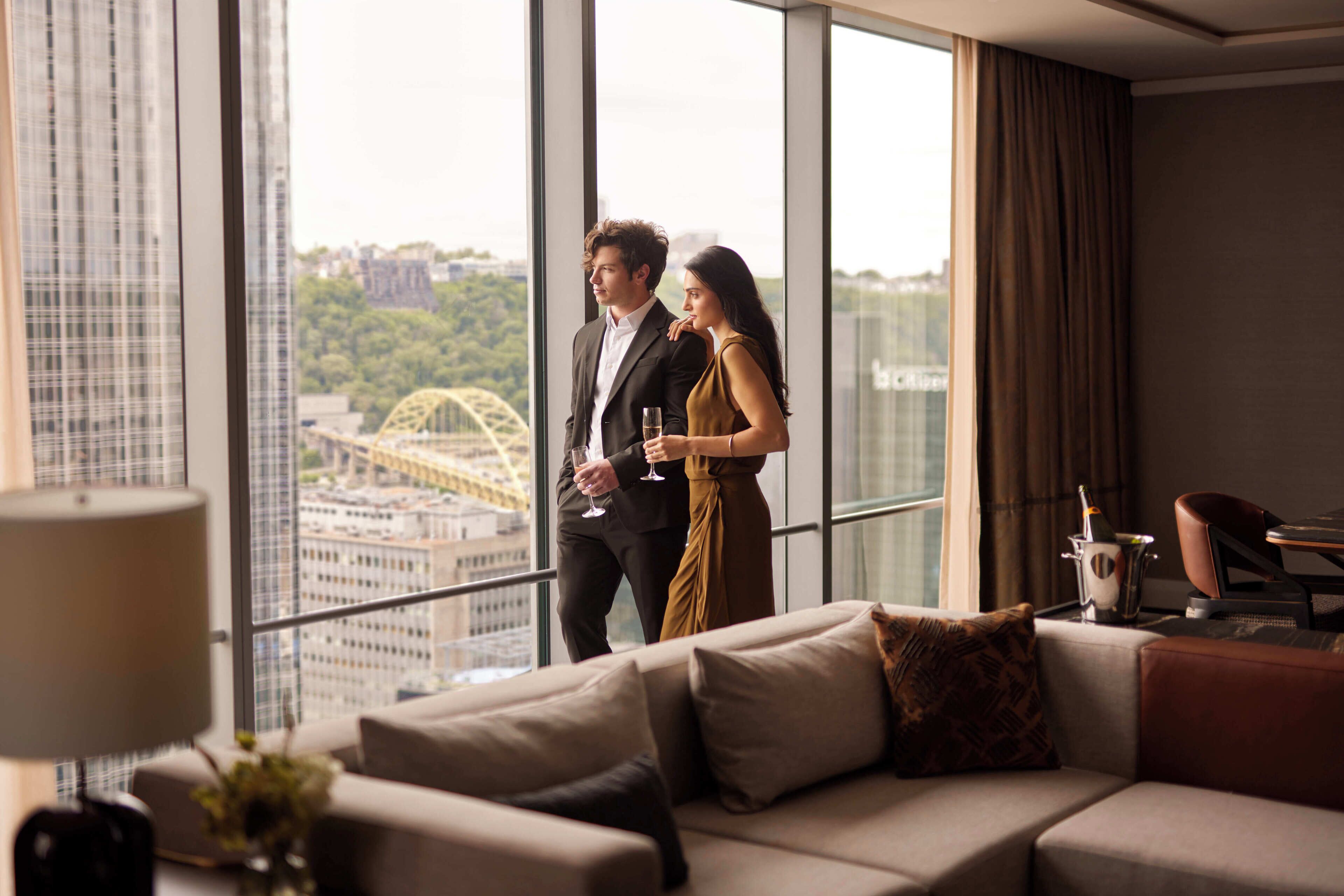 A modern hotel room with a large window, a sofa, and a dining area.