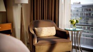A hotel room with a brown armchair, a small table with a vase of flowers, a lamp, and a view of the city through the window. - Fairmont Pittsburgh (Pittsburgh)