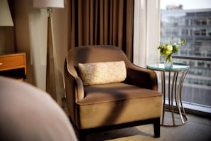 A hotel room with a brown armchair, a small table with a vase of flowers, a lamp, and a view of the city through the window. - Fairmont Pittsburgh (Pittsburgh)