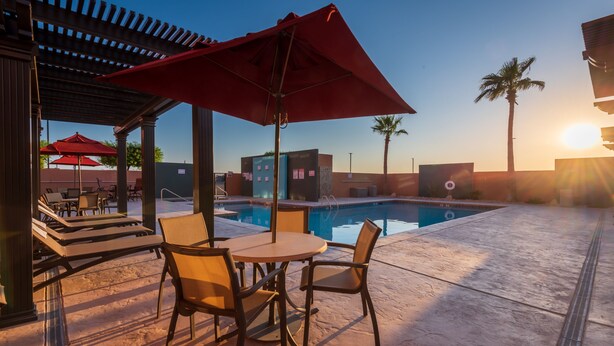 Zona de piscina con sillas de descanso y una mesa bajo una sombrilla roja, con vistas a una piscina.