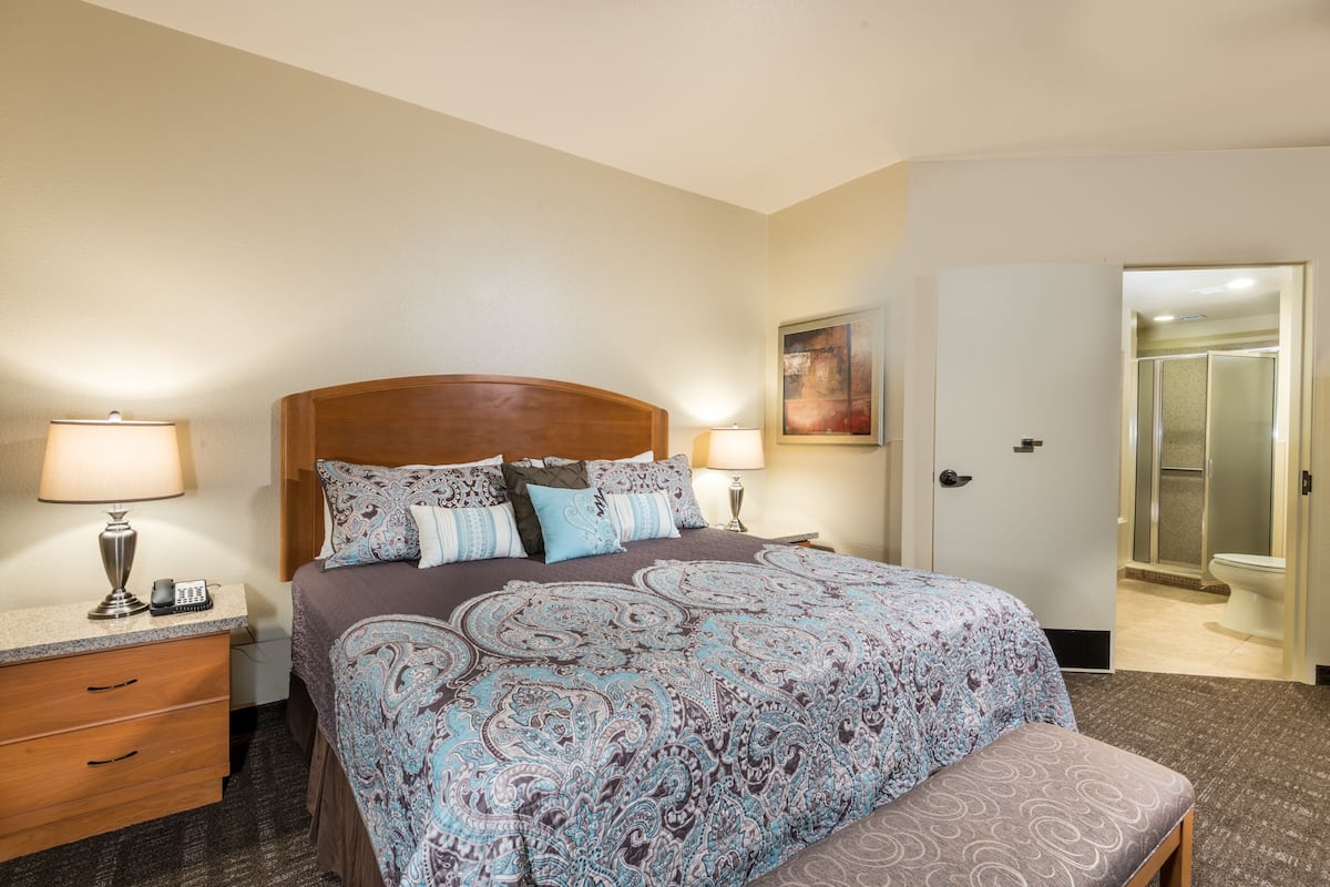 A hotel room with a large bed, bedside lamps, a wooden nightstand, a bathroom with a shower, and a door leading to another room.